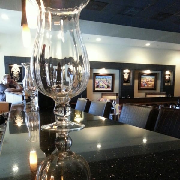 A large glass hurricane lamp on a dark bar counter with seating and Peruvian art in the background.