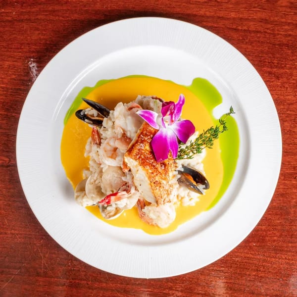 Plated seafood with shrimp, mussels, and seared fish over yellow sauce, garnished with orchid and herbs.