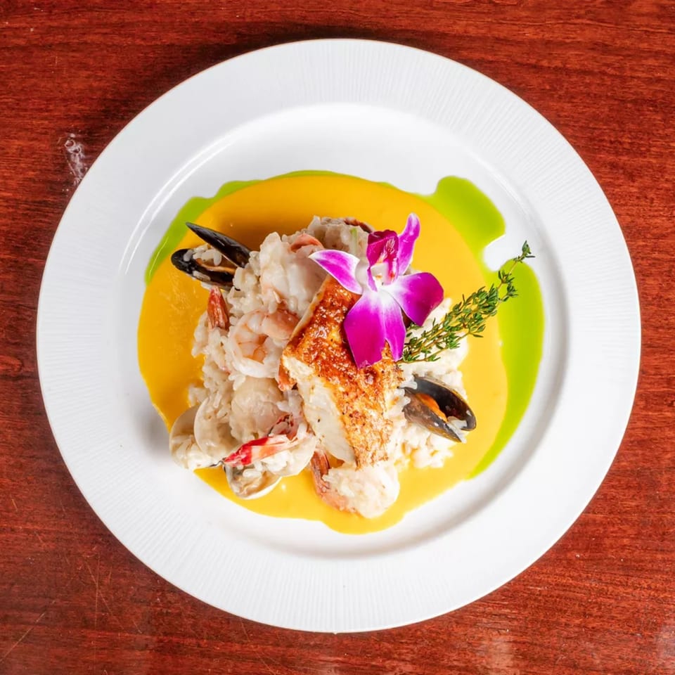 Plated seafood with shrimp, mussels, and seared fish over yellow sauce, garnished with orchid and herbs.
