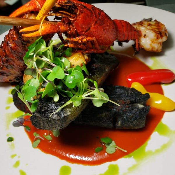 Peruvian dish with a whole red lobster, dark grilled fish, microgreens, and red and yellow sauces.