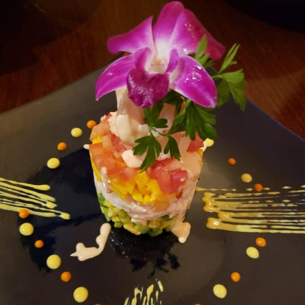 Peruvian ceviche tower with avocado, mango, tomato, and a purple orchid garnish on a black plate.