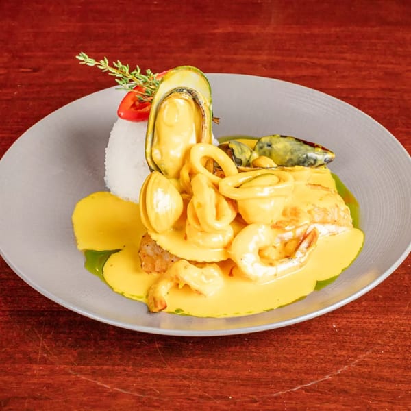 Seafood platter with mussels, shrimp, squid, fish, rice, and creamy yellow sauce garnished with herbs on a gray plate.