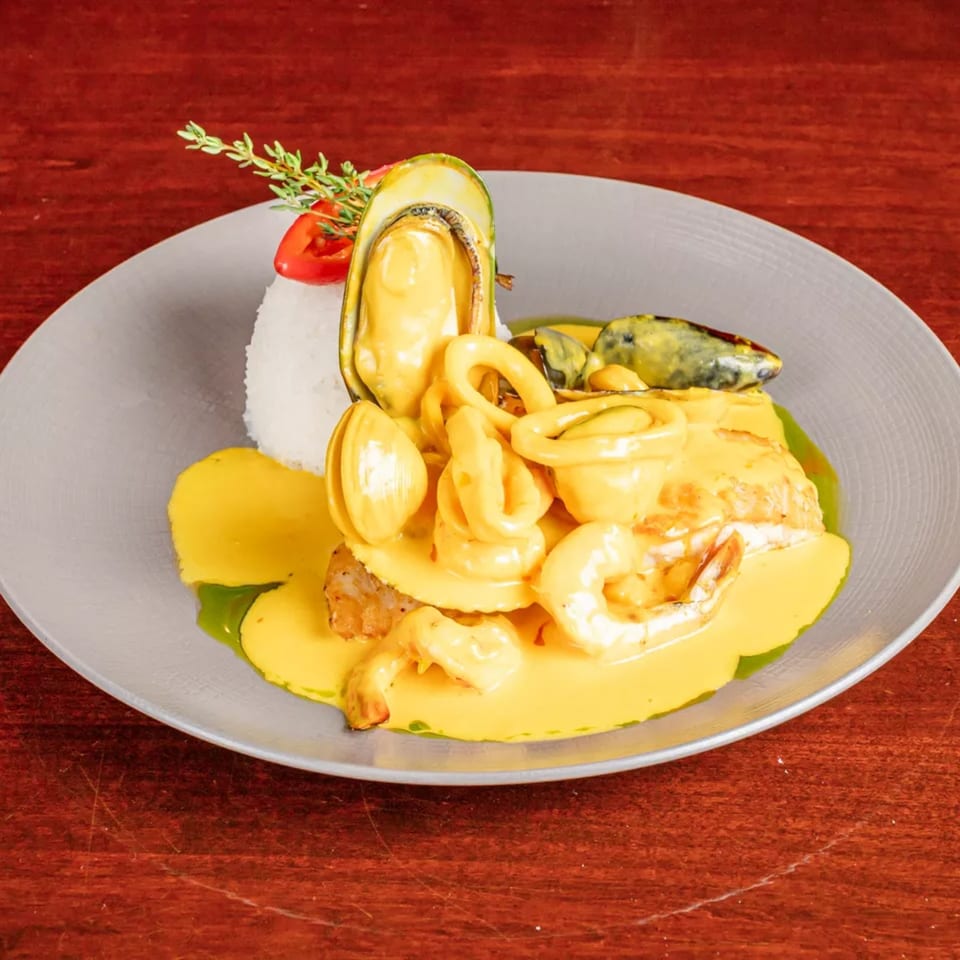 Seafood platter with mussels, shrimp, squid, fish, rice, and creamy yellow sauce garnished with herbs on a gray plate.