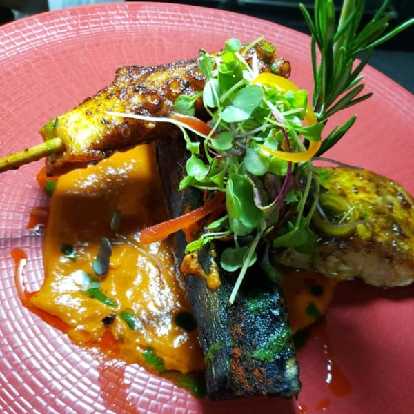 Peruvian dish with grilled octopus, chicken, sweet potato puree, microgreens, and rosemary on a red plate.