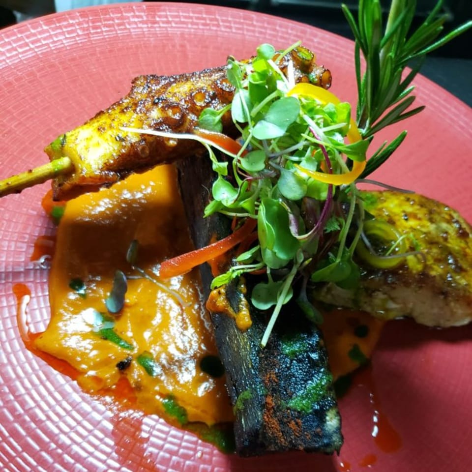 Peruvian dish with grilled octopus, chicken, sweet potato puree, microgreens, and rosemary on a red plate.